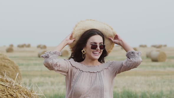 Cheerful Brunette in Sunglasses Comes to Haystack Has Joy and Puts on Hat