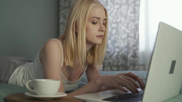 Young Attractive Blonde Woman Works at Home for a Laptop and Drinks Coffee