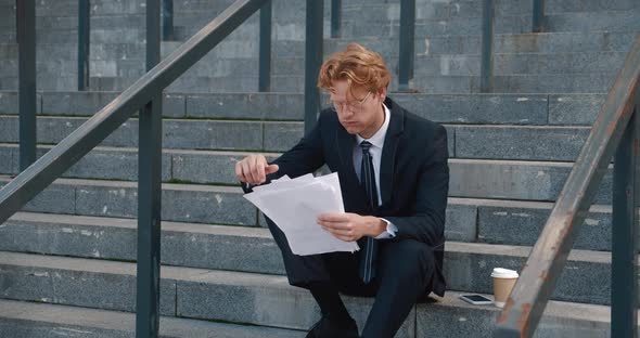 Tired Red Haired Businessman with Business Papers in His Hands Sits on the Steps of an Office