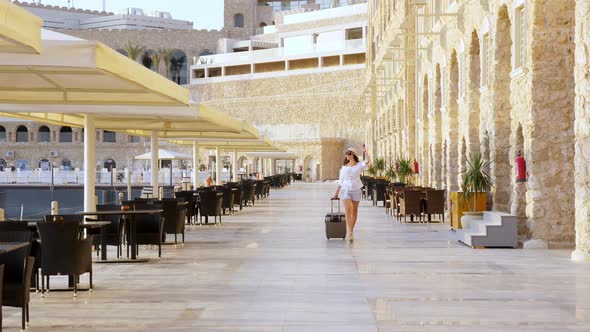 Young Woman in Summer Clothes, Sunglasses and Sun Hat, Walks with Travel Luggage Along a Deserted