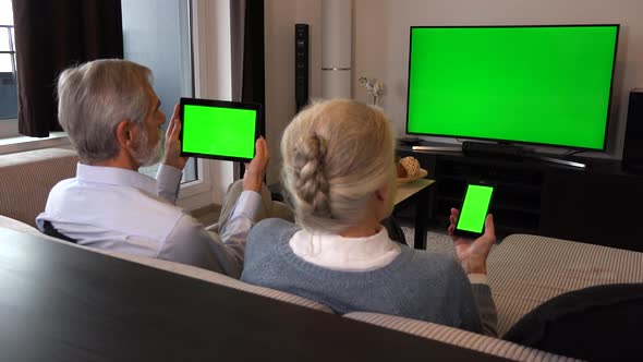 Elderly Couple Sits in A Living Room, Watches Tv with A Green Screen, Looks at Smartphone and Tablet