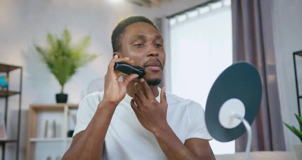 African American which Correcting His Beard wih Trimmer when Sitting in front of Small Mirror