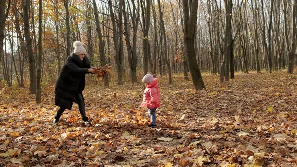 Young woman and her little daughter throw yellow autumn leaves in the city park.