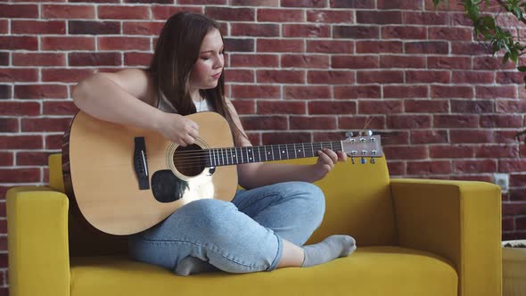 Female Guitarist is Sitting on Sofa in Lotus Position and Playing Guitar Side View