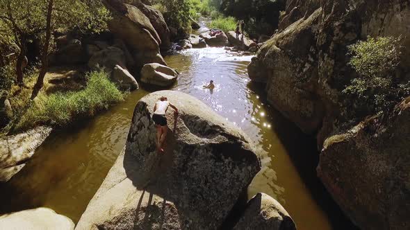 Male Climbing Big Stone in River Raising Tattooed Hands in Front of Friends