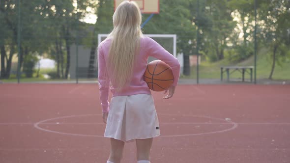 Back View Wide Shot of Sportive Blond Woman Standing with Basket Ball on Outdoor Court. Zoom in To