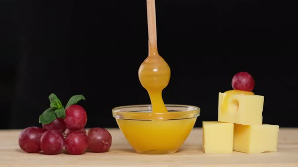 Dips wooden stick to glass bowl with liquid honey. Cheese, Honey and Grapes