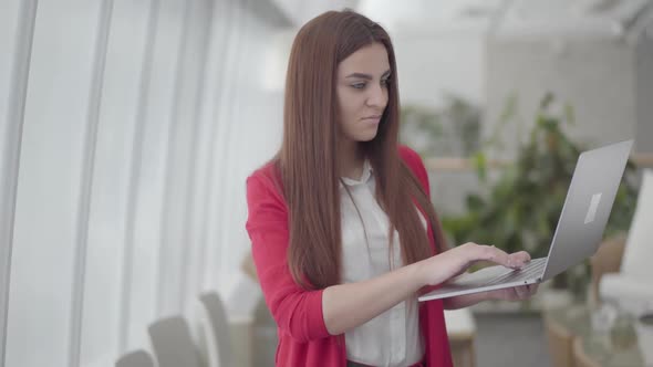 Young Woman in Red Suit Typing on Her Gadget in Modern Light Office. The Girl Closes Her Netbook and