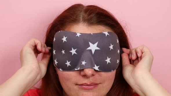 Sleepy Young Caucasian Girl Puts on a Sleep Mask on Her Eyes on a Pink Wall Background