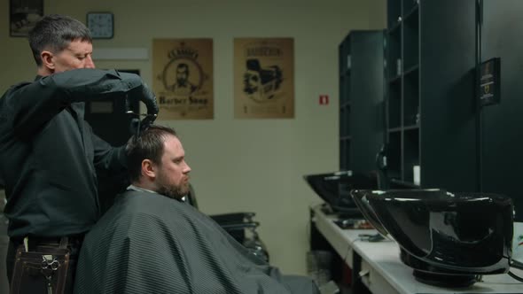 The Hairdresser Makes a Haircut to a Bearded Man with Scissors and a Comb