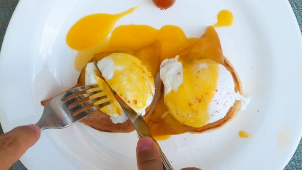 Human Hands Cutting Freshly Cooked Eggs Benedict with Hollandaise Sauce with Fork and Knife