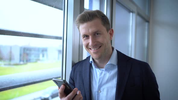 Happy businessman using smartphone, leaning on window in office