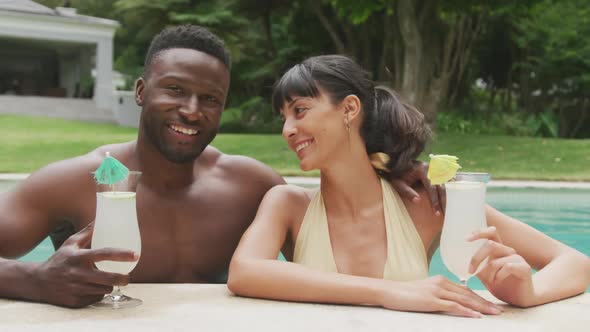 Portrait of happy diverse couple wearing swimming suits and drinking drinks at pool in garden