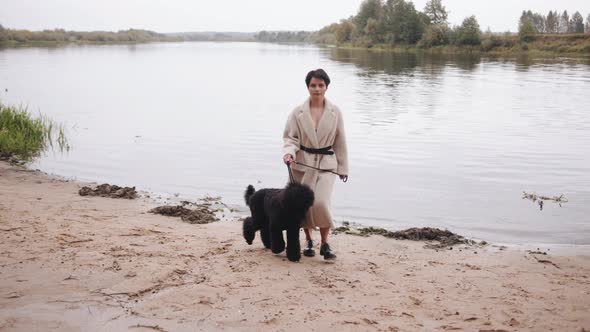 A Young Striking Brunette with Short Hair Goes Along the Sandy Bank of the River and Leads a Large