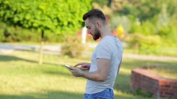 Young Man Standing In The Park With Mobile Phone alt