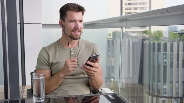 A Young Man Listens To Music on a Smartphone As He Sits on a Balcony in a Luxurious Apartment