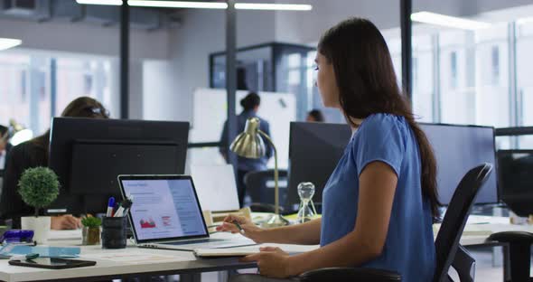 Caucasian businesswoman sitting at desk using laptop and writing notes in notebook in busy office