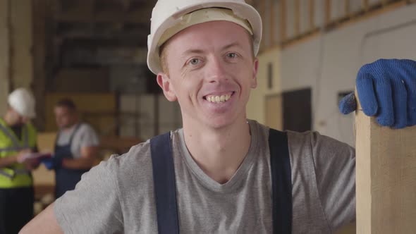 Portrait of a Young Caucasian Builder in White Helmet Standing with Wooden Plank and Smiling