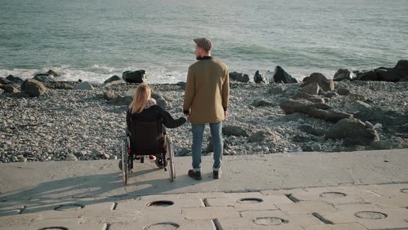 Wheelchair Woman User and Healthy Man Are Holding Hands and Watching Seascape