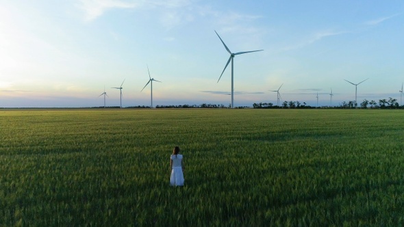 Beautiful girl standing on a green wheat field with windmills for electric power production