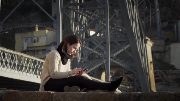 Gorgeous Lady Using Smartphone While Sitting By The Steel Bridge Structure In Porto, Portugal - full