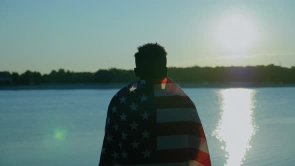 Afroamerican Man with American Flag on Shoulders Looks Into Distance at Sunrise