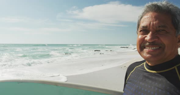 Portrait of happy senior hispanic man walking on beach with surfboard