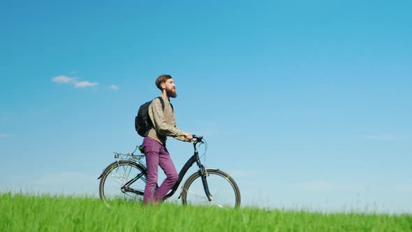 A Young Hipster Man Drives a Bicycle Through a Green Meadow