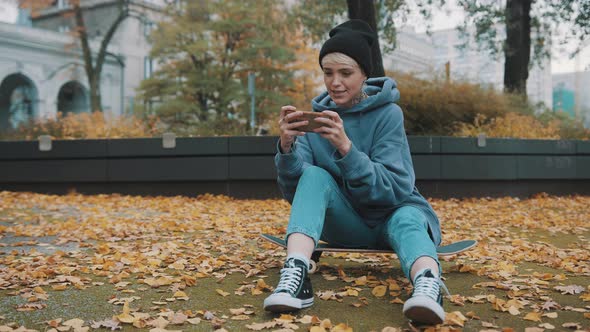 Young Hipster Woman Sitting on the Skateboard and Playing Games on Her Phone in the Park in Outumn