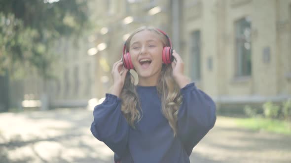 Portrait of Joyful Little Girl Walking Along Sunny Schoolyard Listening To Music in Headphones