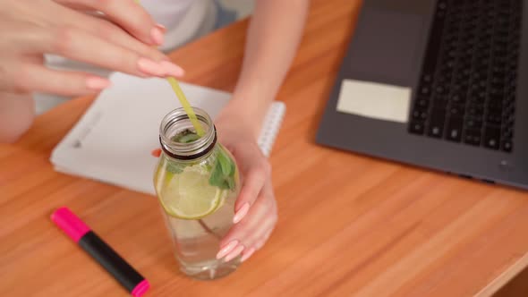 Blonde Girl at the Table in the Kitchen Drinking Lemonade From a Straw Working at a Laptop