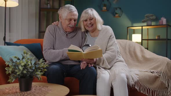 Senior Grandparents Couple Relaxing Reading Book Talking Enjoying Leisure Hobbies at Night Home