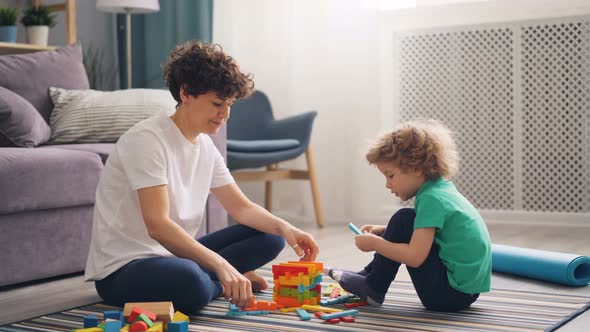 Happy Child Boy Playing with Wooden Toys While Mother Is Helping Him in House