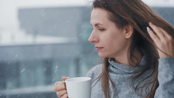 Caucasian Woman Stays on Balcony During Snowfall with Cup of Hot Coffee or Tea