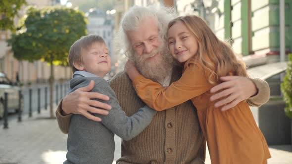 Portrait of Smiling Grey-haired Senior Man Hugging Children Running To Grandfather. Camera