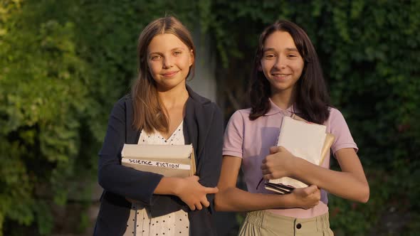 Two Positive Teenage Girls Standing in Golden Sunshine in Park with Books Looking at Camera Smiling