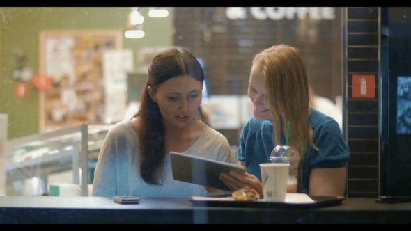 Female Friends Talking And Using Touch Pad In Cafe alt