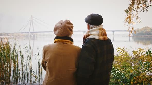 Elderly Husband Hugging Wife Smiling and Talking to Her Standing on Bank of a River and Looking at