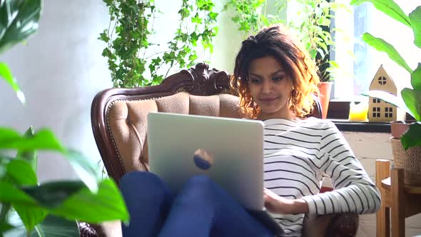 Woman Uses Laptop and Sits in Chair on Background of Green Plants at Home Spbd