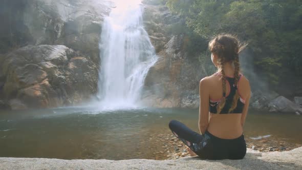 Blond Woman Sits on Stone Watches Foamy Waterfall