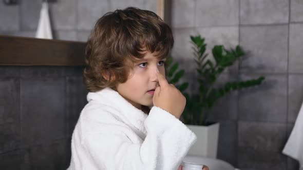 Little Boy in white Robe with Dark Curly Hair Applies Cream on Nose