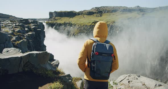 Man in Yellow Jacket and Backpack Walking on Cliff Edge Enjoying Detifoss Waterfall in Iceland