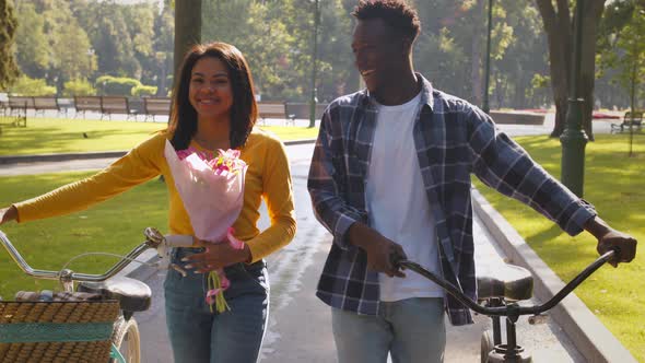 Lovely Young African American Man and Woman Walking with Bicycles Through Public Park Follow Shot