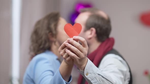 Closeup of Red Paper Heart in Male and Female Hands with Blurred Adult Couple Kissing at Background