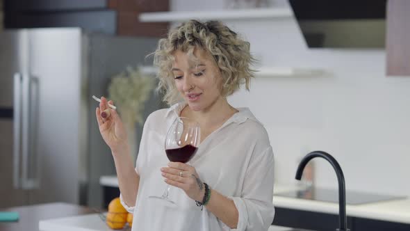 Medium Shot of Carefree Smiling Happy Young Woman Smoking Cigarette Drinking Red Wine at Home