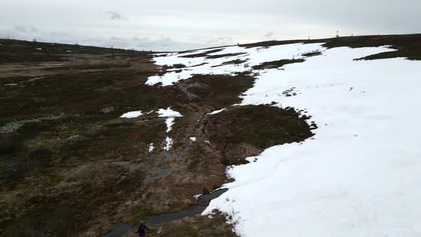 A woman and her dog walking on a mountain path with snow on their right hand side. Aerial reverse re