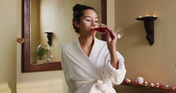 Relaxed biracial woman with vitiligo wearing bathrobe and drinking wine in bathroom