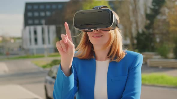 A Happy Woman in the Suit is Standing Outside and Gestures in Augmented Reality in 3D VR Goggles