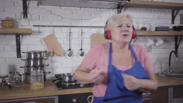 Funny Caucasian Old Woman in Headphones Singing and Dancing at the Kitchen. Mature Cheerful Retiree