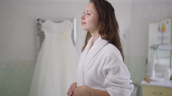Gorgeous Happy Young Bride Admiring White Wedding Dress Hanging in Bathroom Smiling Dreaming